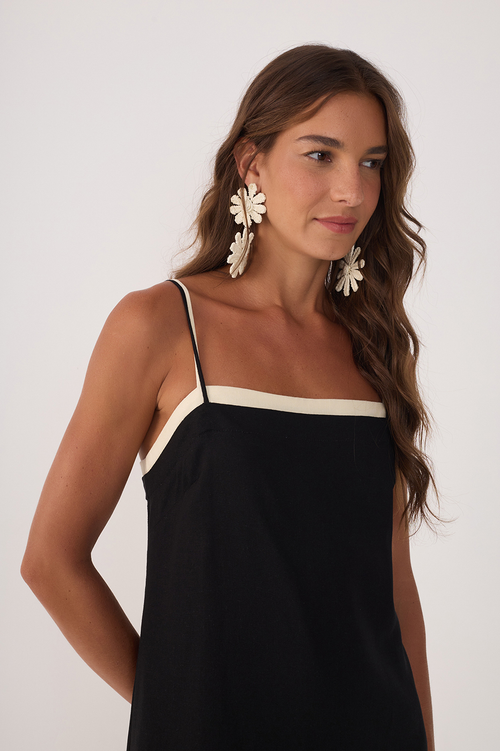 Woman wearing a knee length black dress with white layered trim and white flower statement earrings against a white background