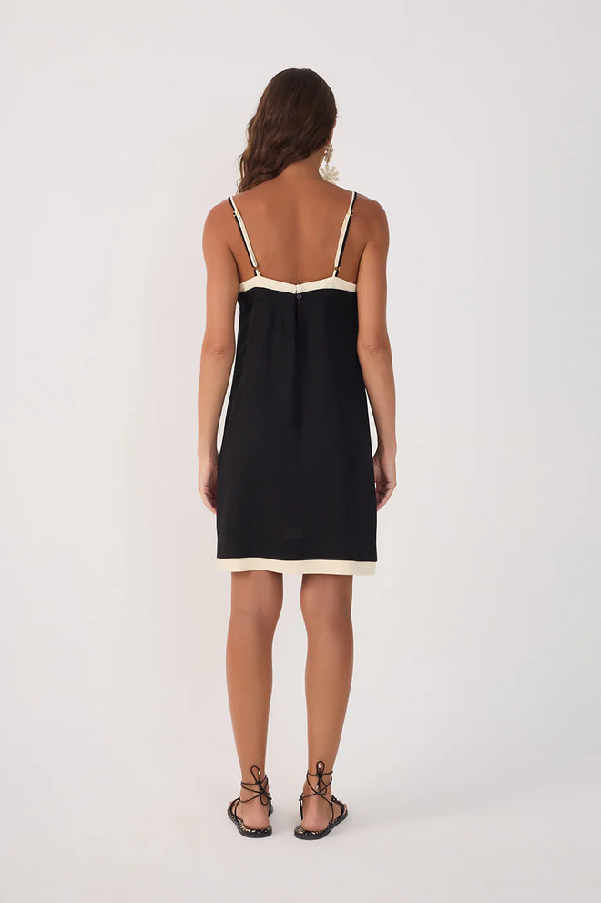 Back view of a woman wearing a knee length black dress with white layered trim and white flower statement earrings with black strappy sandals against a beige background
