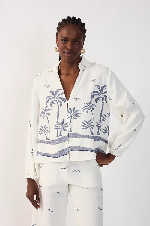 Woman wearing a collared white shirt with blue palm tree pattern and white printed pants on a plain background