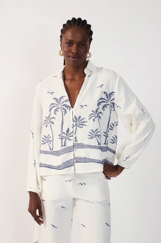 Woman wearing a collared white shirt with blue palm tree pattern and white printed pants on a plain background