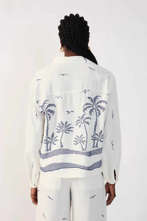 Woman wearing a collared white shirt with blue palm tree pattern and white printed pants on a plain background