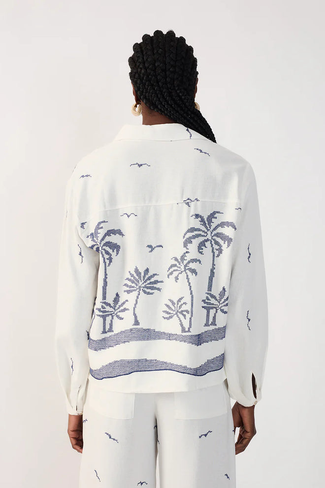 Woman wearing a collared white shirt with blue palm tree pattern and white printed pants on a plain background