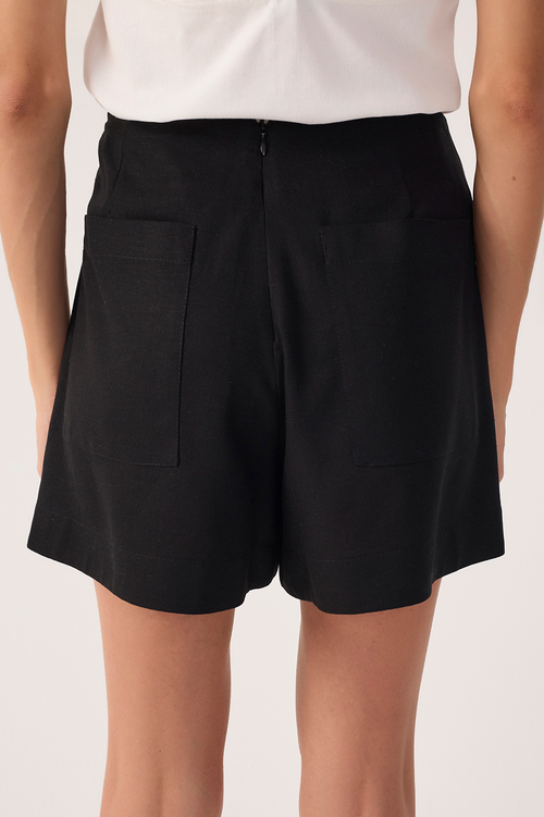 Close up back view of a woman wearing a white top and black shorts with pockets on a white background