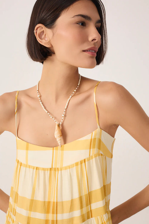 Woman wearing a yellow and white checkered dress with a seashell necklace on a plain background