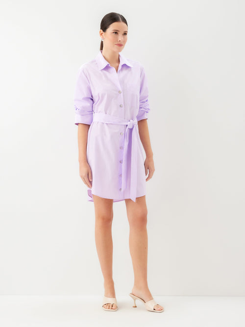 Side view of a woman wearing a short light purple dress with a belt and kitten heels on a plain background