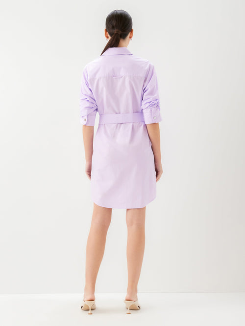 Back view of a woman wearing a short light purple dress with a belt and kitten heels on a white background