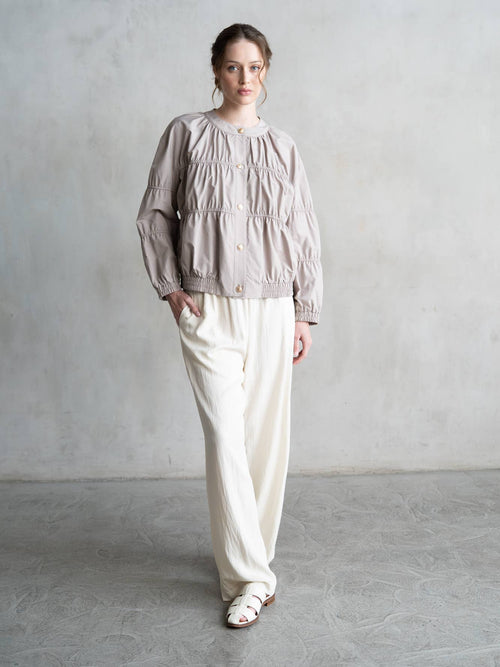 Brown hair blue eyed woman wearing a grey jacket with snap button closure and ruching over white linen pants and white sandals against a plain background