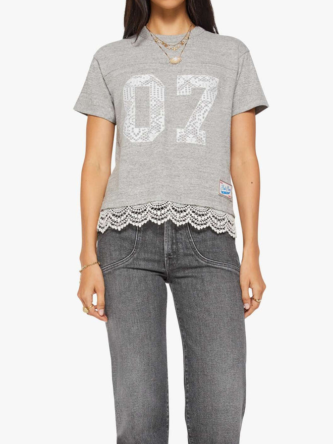 Woman wearing a gray football jersey t-shirt with '07' and lace details, paired with gray jeans on a white background.