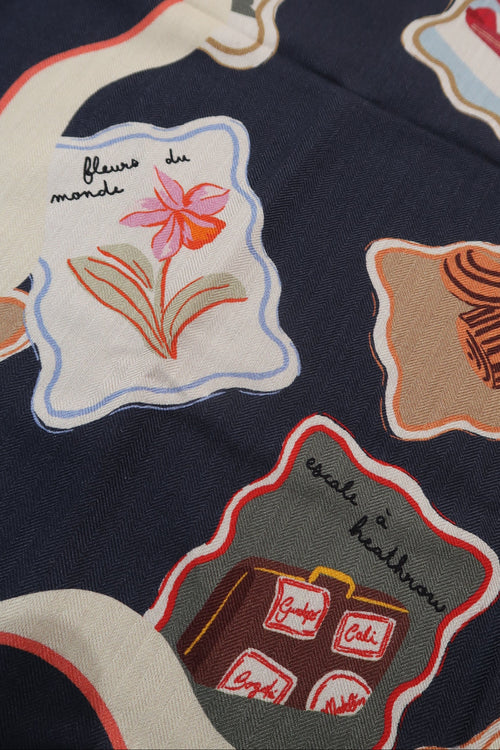 Close-up of a navy blue fabric with a whimsical patchwork print featuring illustrated motifs like a pink flower, a striped hat, a suitcase, and handwritten text labels.