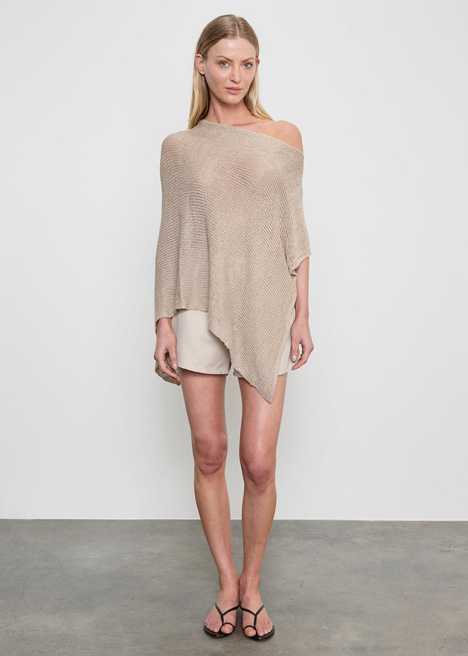 Woman wearing a beige off-shoulder poncho top, black sandals and shorts on a plain background