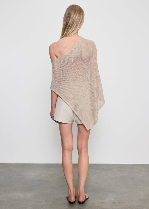 Woman wearing a beige off-shoulder poncho top, black sandals and shorts on a plain background