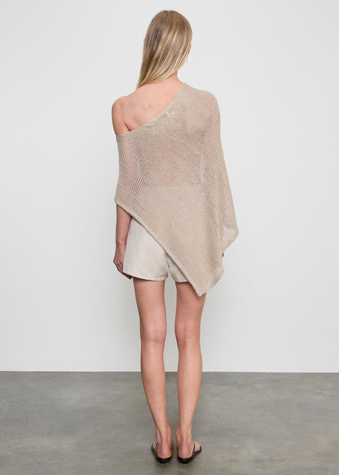 Woman wearing a beige off-shoulder poncho top, black sandals and shorts on a plain background