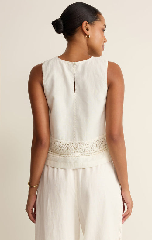 Back view of a woman wearing a white sleeveless top with lace crochet details with white pants on a beige background