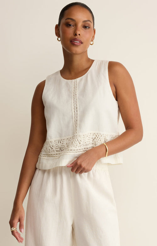 Woman wearing a white sleeveless top with lace crochet details with white pants on a beige background