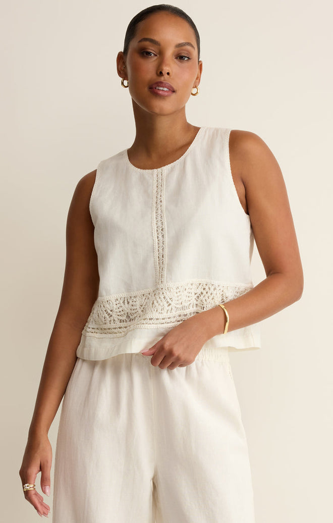 Woman wearing a white sleeveless top with lace crochet details with white pants on a beige background