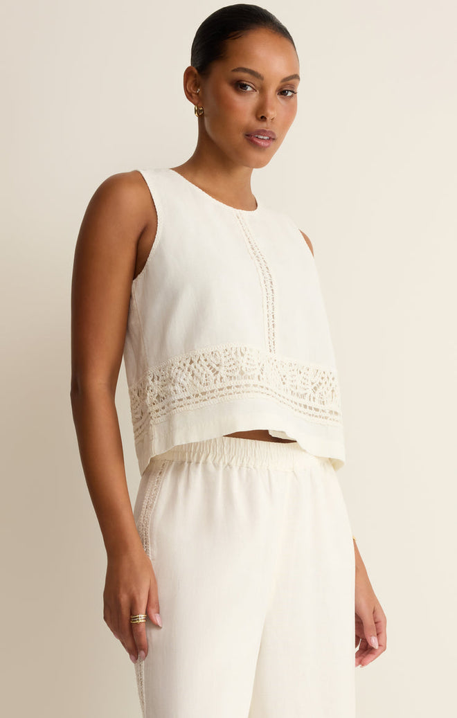 Woman wearing a white sleeveless top with lace crochet details with white pants on a beige background