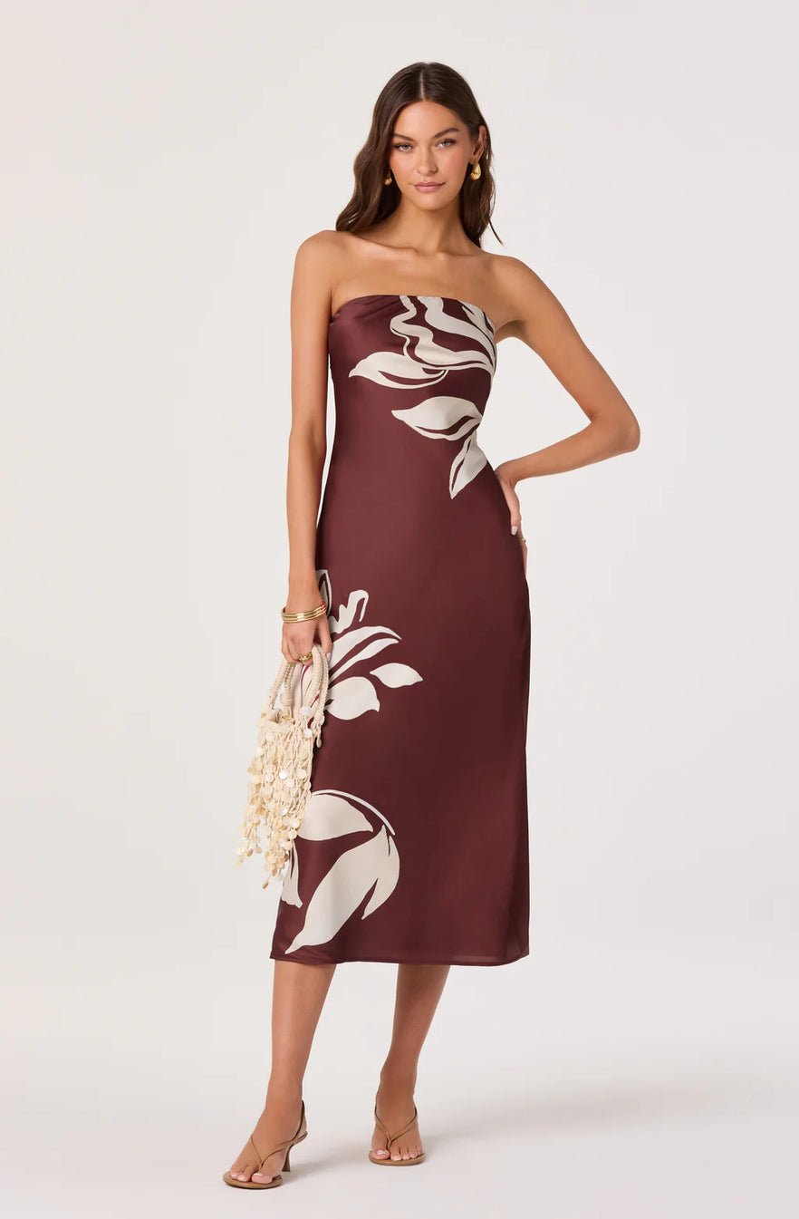 A woman models a strapless midi dress in a deep burgundy color with a large white floral print. She is holding a small woven clutch and wearing gold jewelry and nude heels, set against a plain light gray studio background.