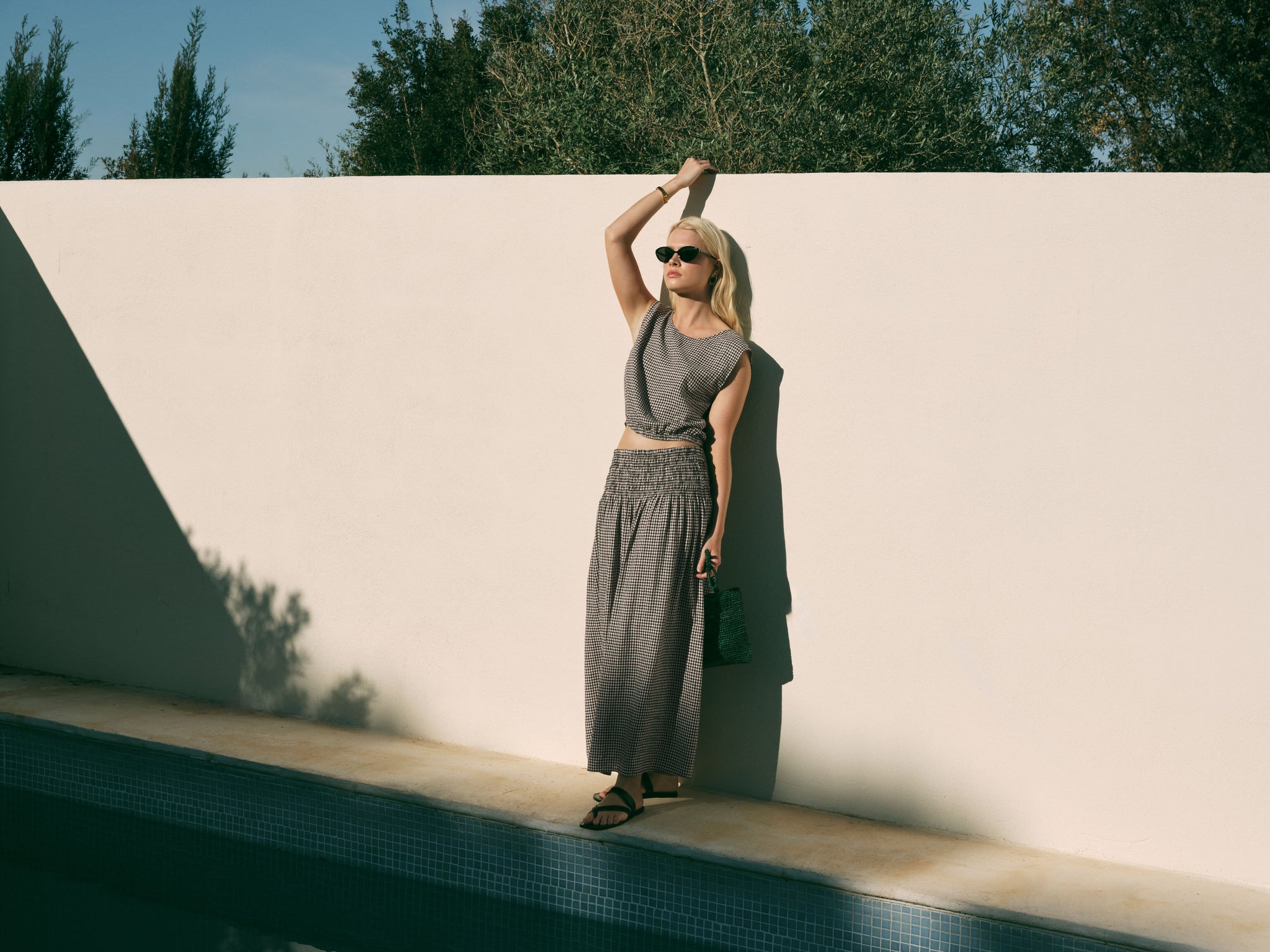 A woman wearing a matching gray two-piece set consisting of a sleeveless crop top and a long smocked maxi skirt, standing next to a pool against a minimalist beige wall.